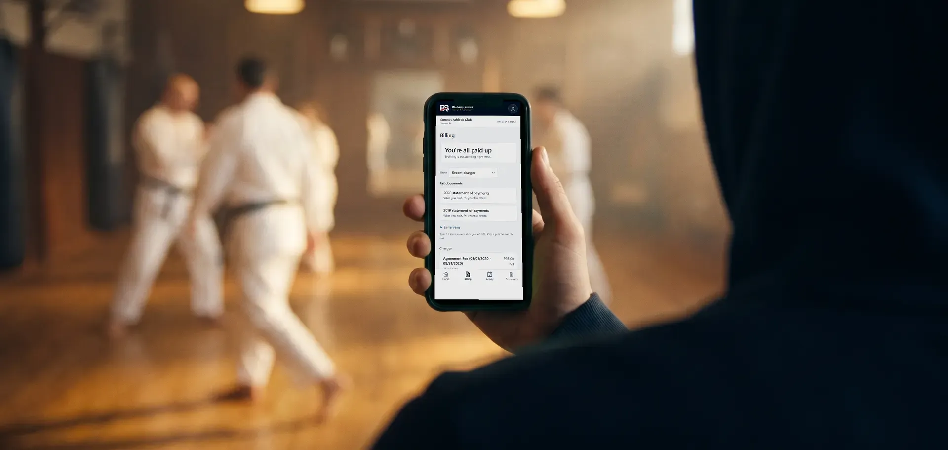 A parent at the side of a martial arts class holding a phone showing the member portal Billing screen: You're all paid up, with statements of payments.